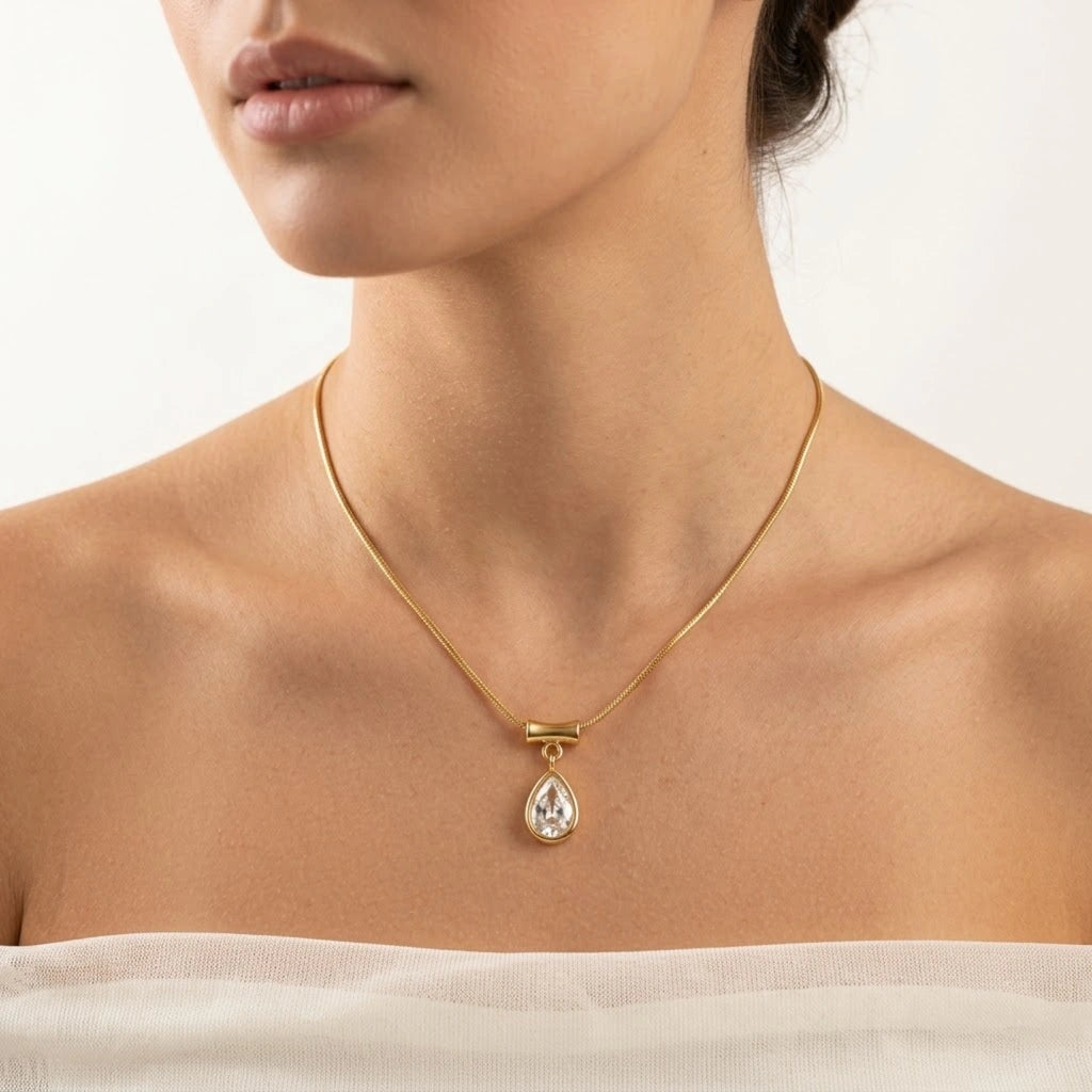 A close-up of a model wearing a delicate gold chain necklace with a teardrop-shaped clear gemstone pendant, styled against a clean white background.